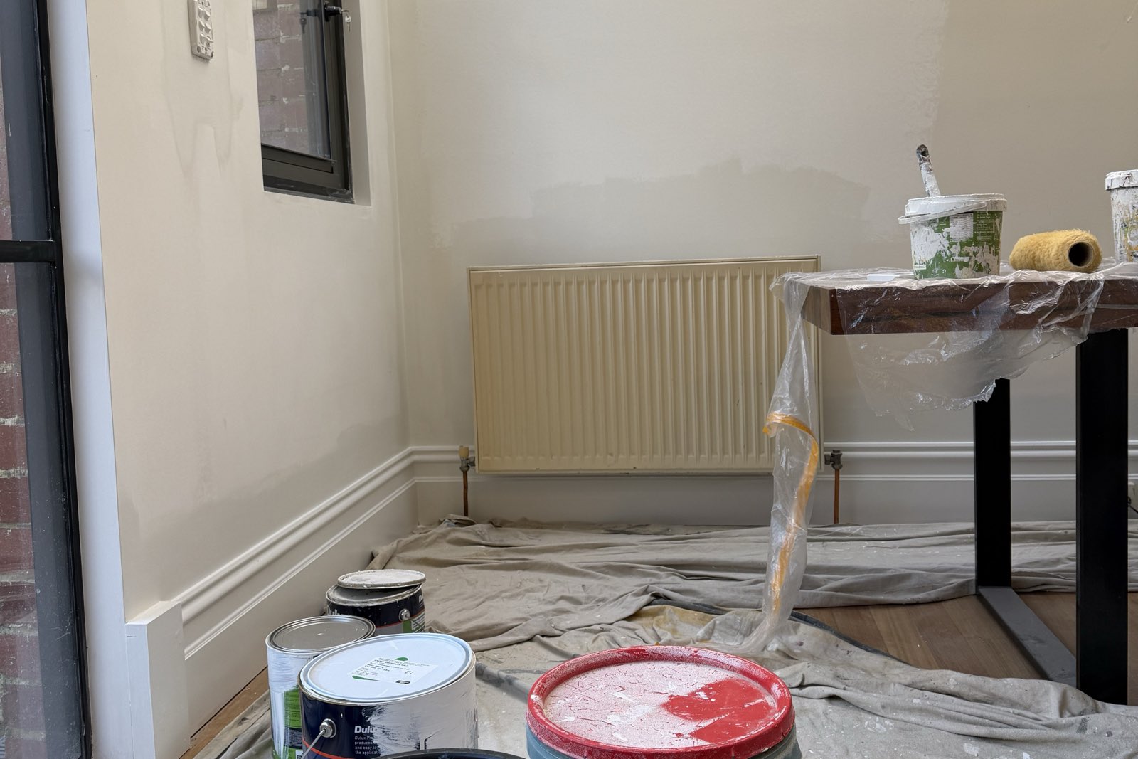 A room part-way through painting: drop sheets over the floor, a covered trestle, open paint tins, a radiator and skirting boards masked, and a wall in an unfinished state.