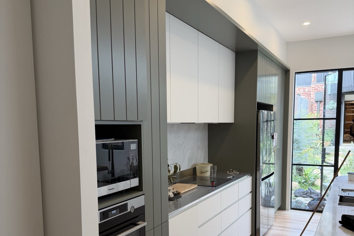 A galley kitchen with deep green vertical panelling, white overhead cabinets, integrated ovens and a pale benchtop, beside a black-framed glazed door.