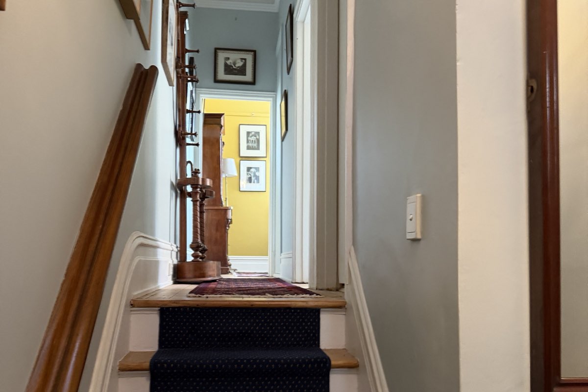 A staircase and hallway with pale grey-green walls, white-painted stringers and skirting, a polished timber handrail and a yellow wall at the far end.