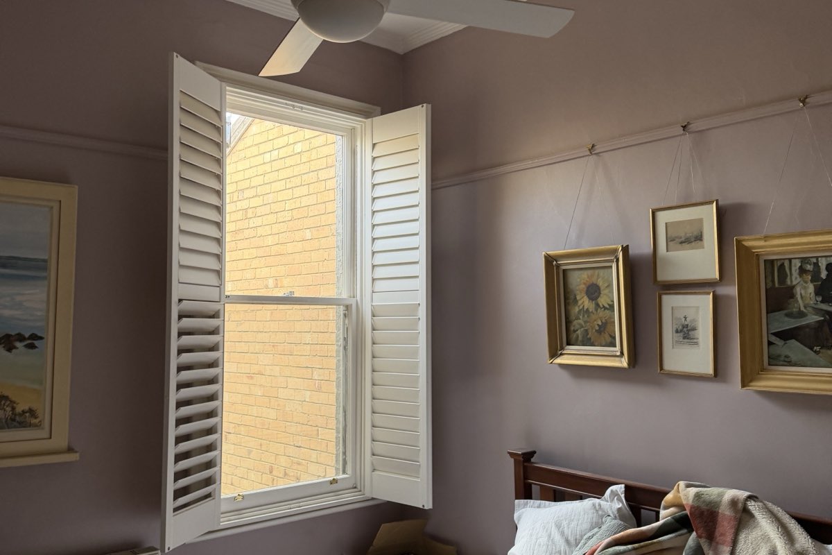 A bedroom painted in soft mauve, with a white-painted window frame, plantation shutters, picture rail and framed pictures.