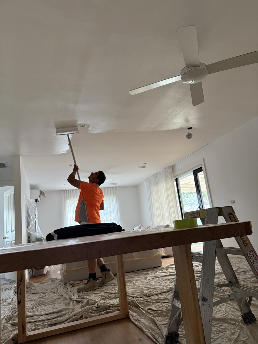 A painter using an extension roller to paint a white ceiling, with drop sheets and covered furniture in the room.