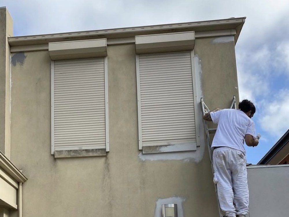 An upper exterior facade during preparation, with patching around shutters and a painter working from a ladder.