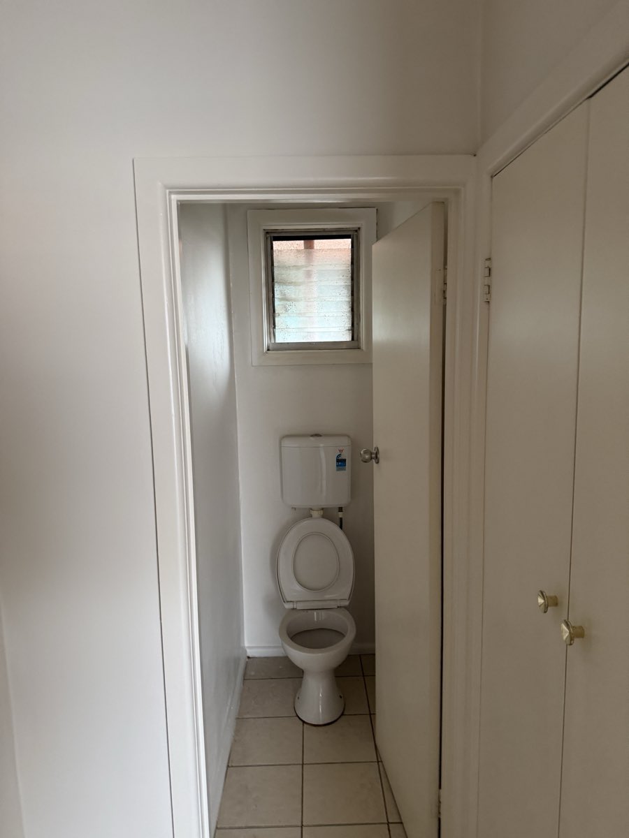 A small toilet room has light painted walls, a high window and a tiled floor.