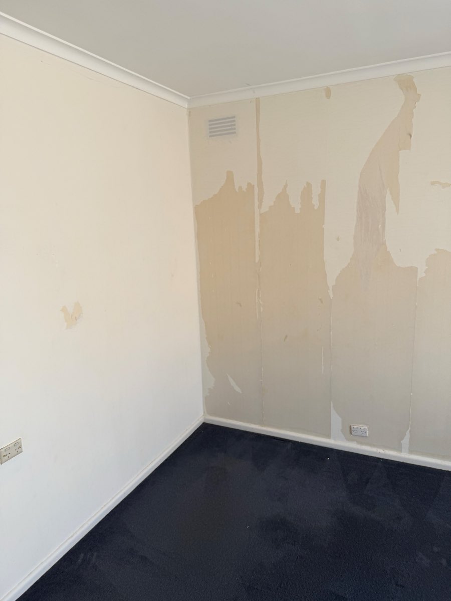 Interior wall with several broad scraped and filled patches, a vent near the ceiling and dark carpet below.