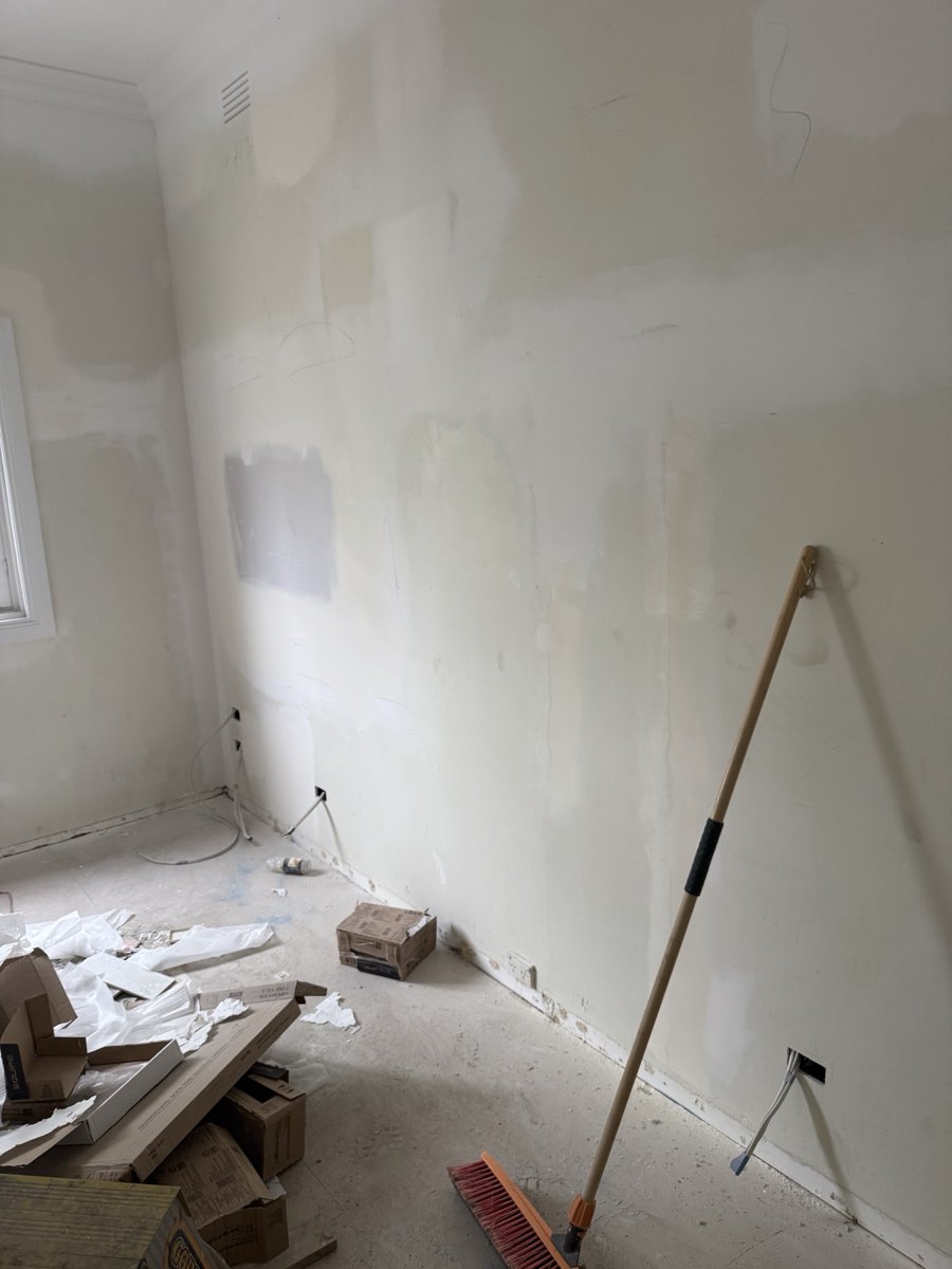 An interior wall with several rectangular patched and filled sections, in a stripped-back room with flooring materials, boxes and a broom on a bare concrete floor.