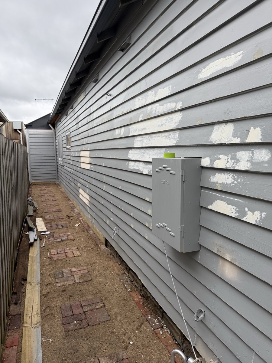 A grey-painted weatherboard side wall with several lighter patch marks over prepared sections, and a wall-mounted electrical switchboard.